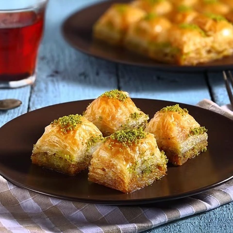 Four pieces of baklava with pistachio on a dark plate, with tea and more baklava in the background.