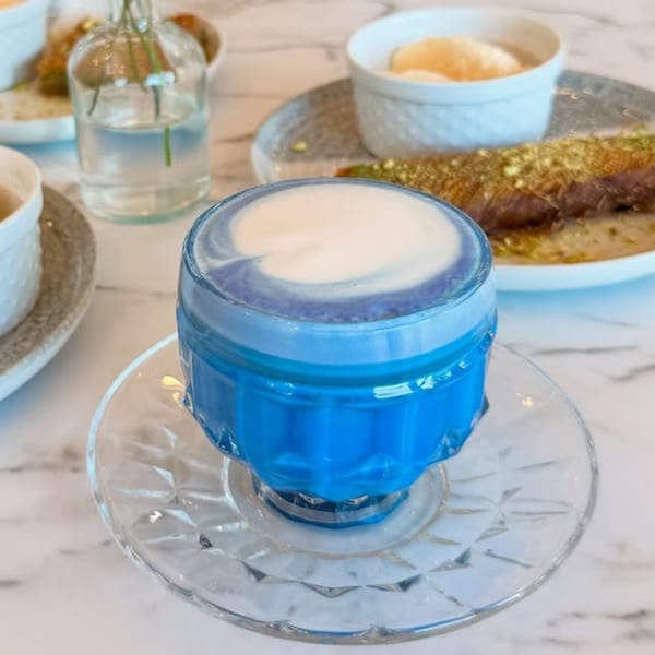 A vibrant blue drink with white foam in a clear glass on a marble table, with desserts in the background.
