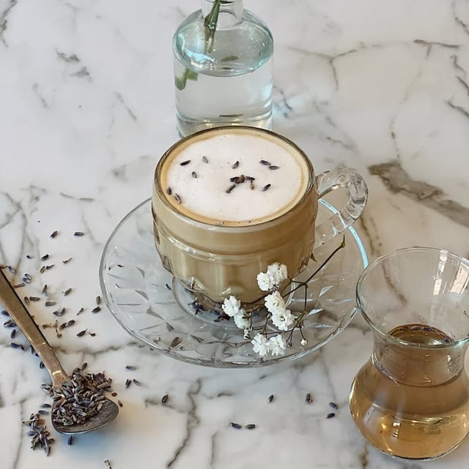 A lavender latte with foam and lavender buds, a spoon of lavender, and a small vase on marble.